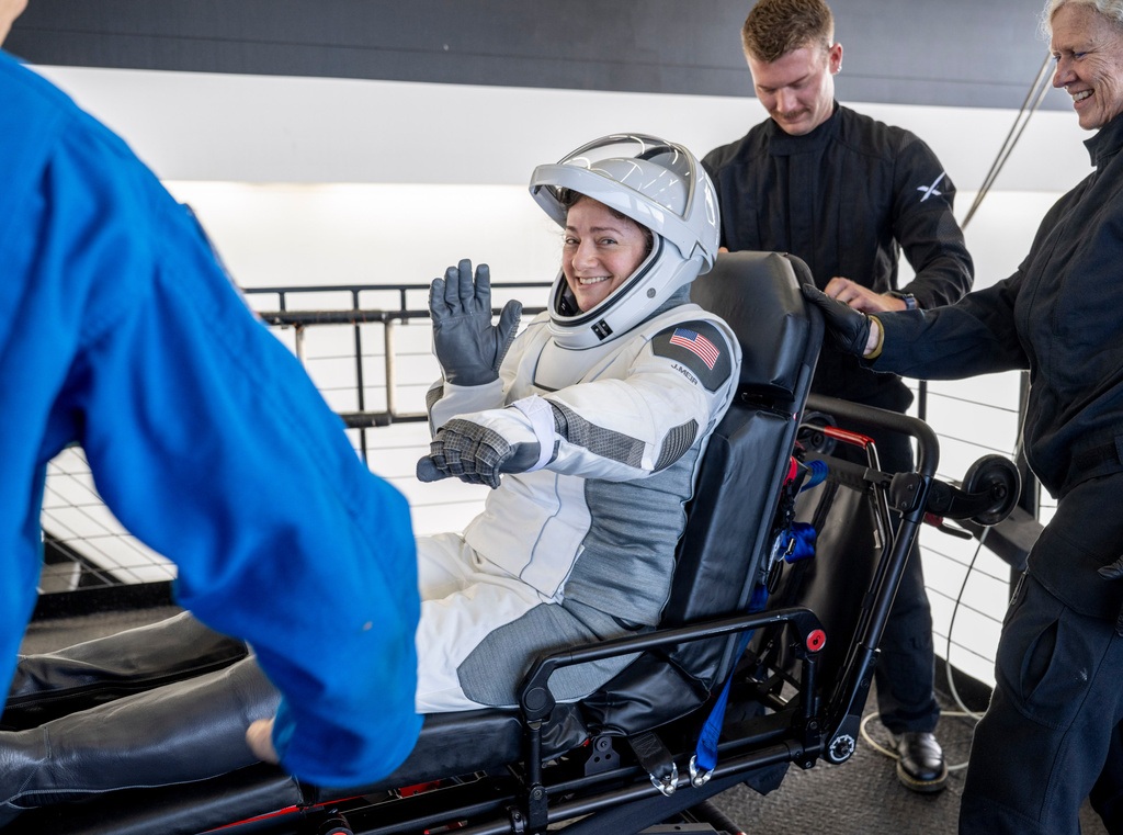 Astronaut in white SpaceX suit waving while seated in spacecraft with crew members assisting