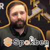 Man wearing Sparkbee branded black polo shirt at promotional event with gold and black backdrop