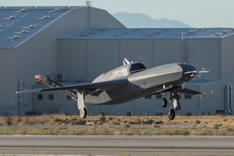 Silver and black experimental aircraft landing near industrial hangar with mountains in background