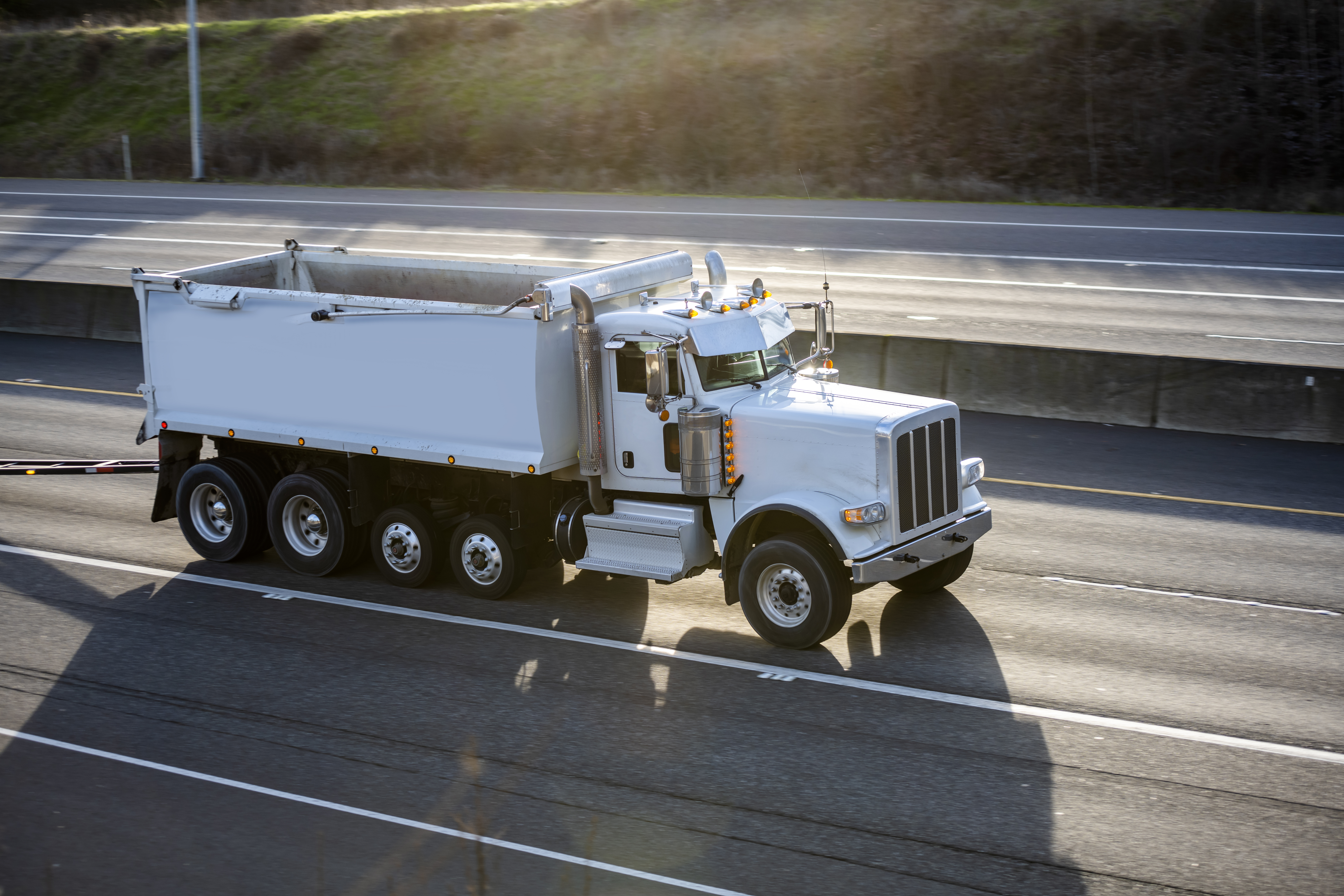 White Peterbilt dump truck traveling on a multi-lane highway with grassy hillside in background