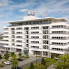 Modern white multi-story commercial building with glass windows and flags on rooftop, surrounded by landscaping and parked vehicles