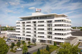 Modern white multi-story commercial building with glass windows and flags on rooftop, surrounded by landscaping and parked vehicles